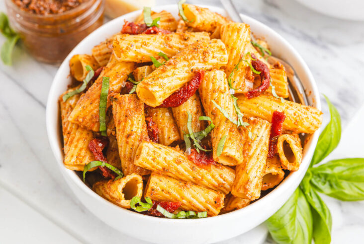 A white bowl containing cooked rigatoni pasta coated in sundried tomato pesto garnished with fresh basil.