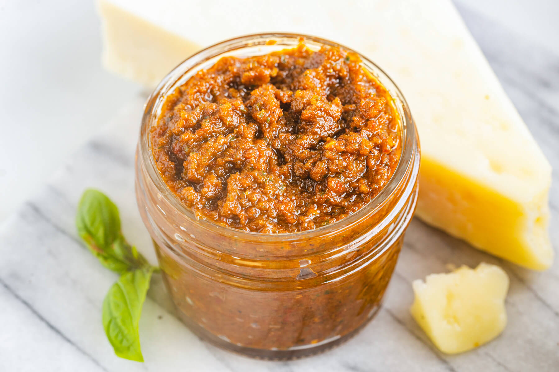 A jar of vibrant Sundried Tomato Pesto surrounded by fresh basil, Parmesan cheese, and garlic.