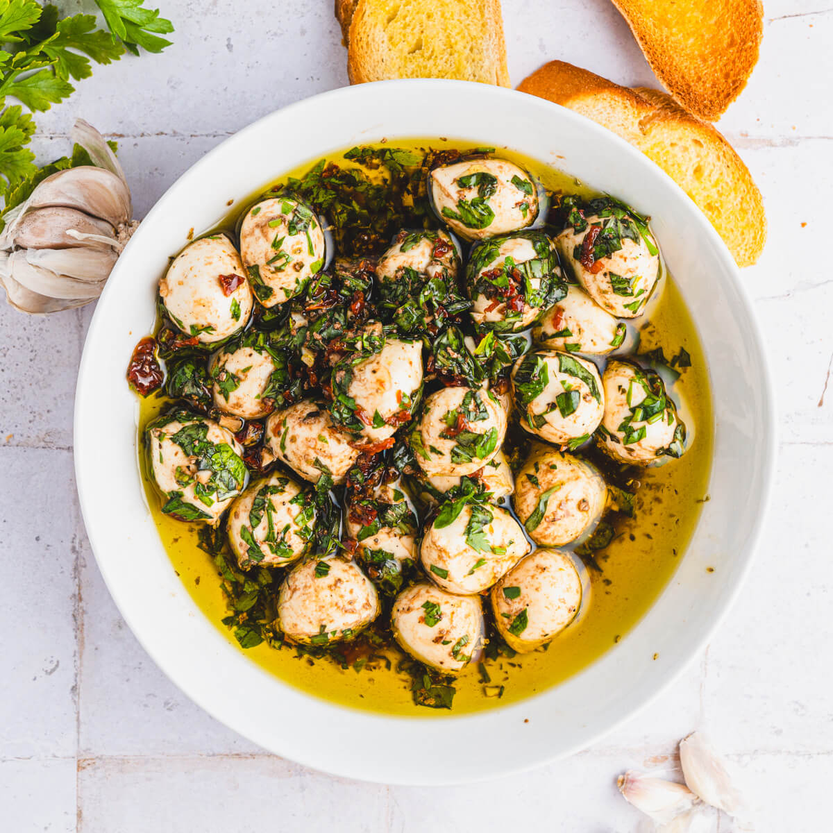 Two golden toasts beside a white bowl filled with bite sized mozzarella balls coated in vibrant green herbs, yellow olive oil, and dotted with sundried tomatoes.