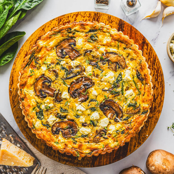 A whole Spinach Mushroom Quiche in a golden baked crust on a round wooden serving board.