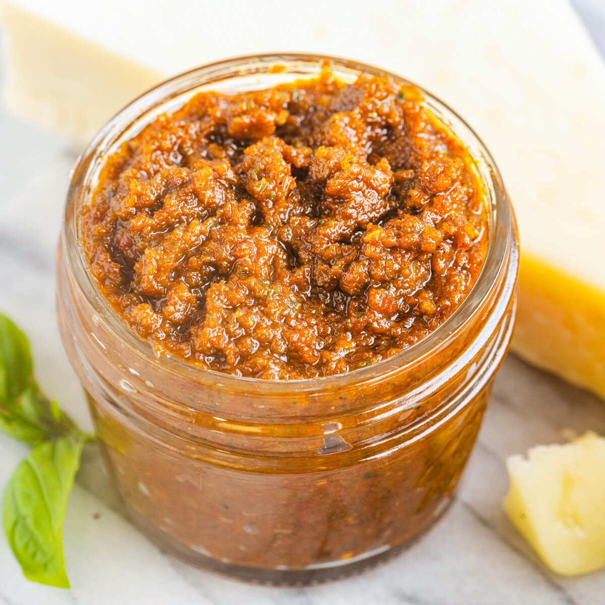 A jar of vibrant Sundried Tomato Pesto surrounded by fresh basil, Parmesan cheese, and garlic.