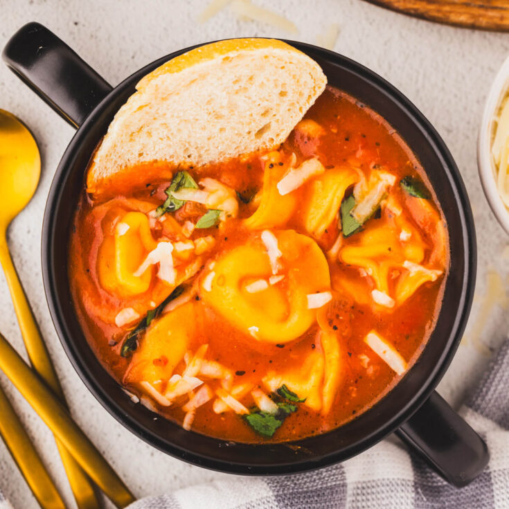 A black soup bowl filled with cooked tortellini pasta in a creamy tomato broth with a side of bread.