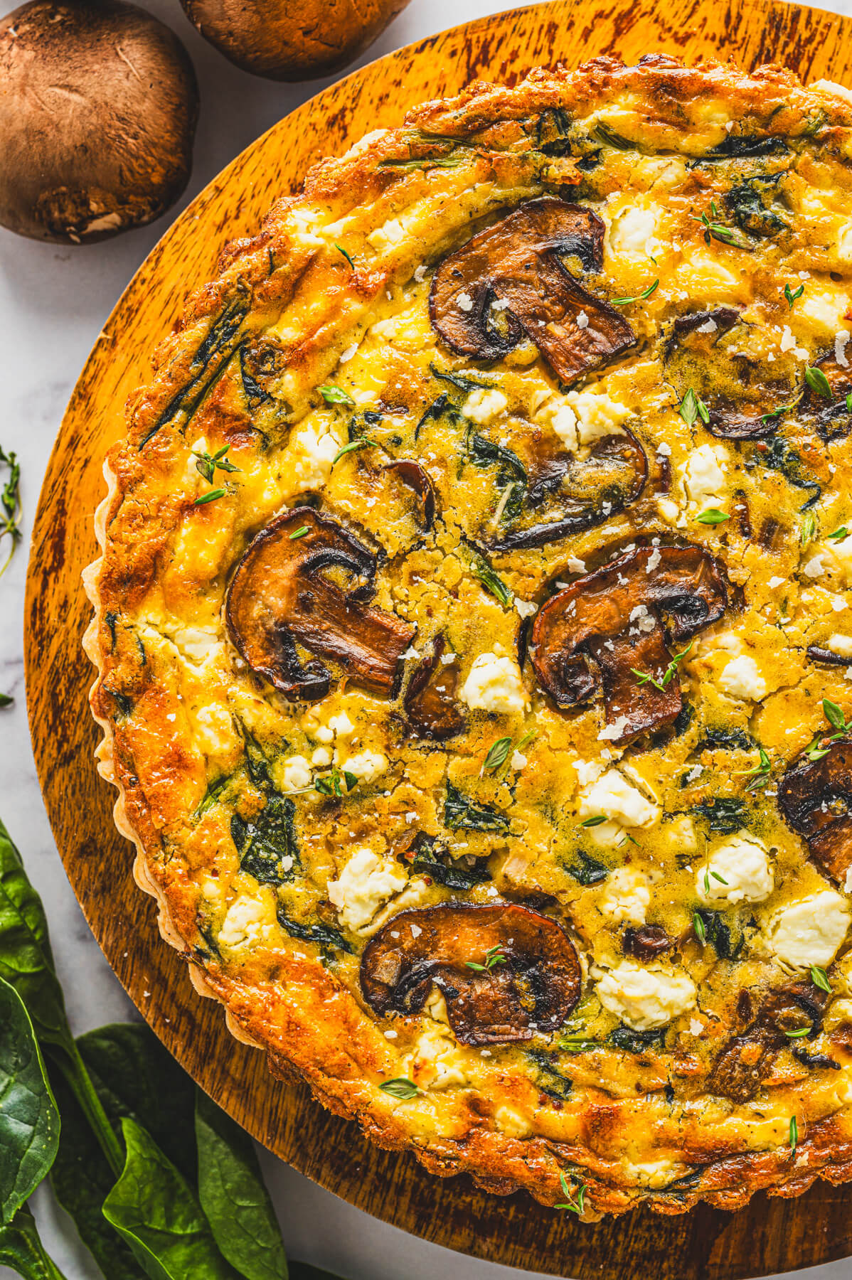 Half of a Spinach Mushroom Quiche in a golden baked crust on a round wooden serving board.