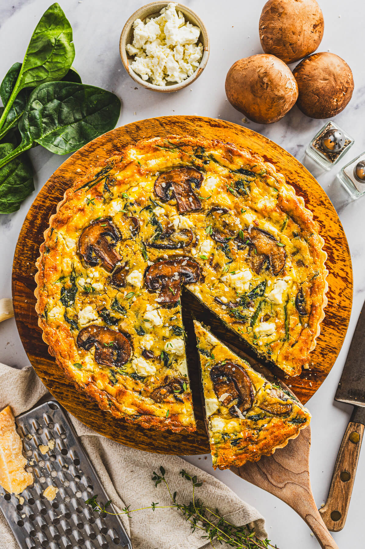 A slice of Spinach Mushroom Quiche in a golden baked crust on a wooden spatula resting above the whole quiche.