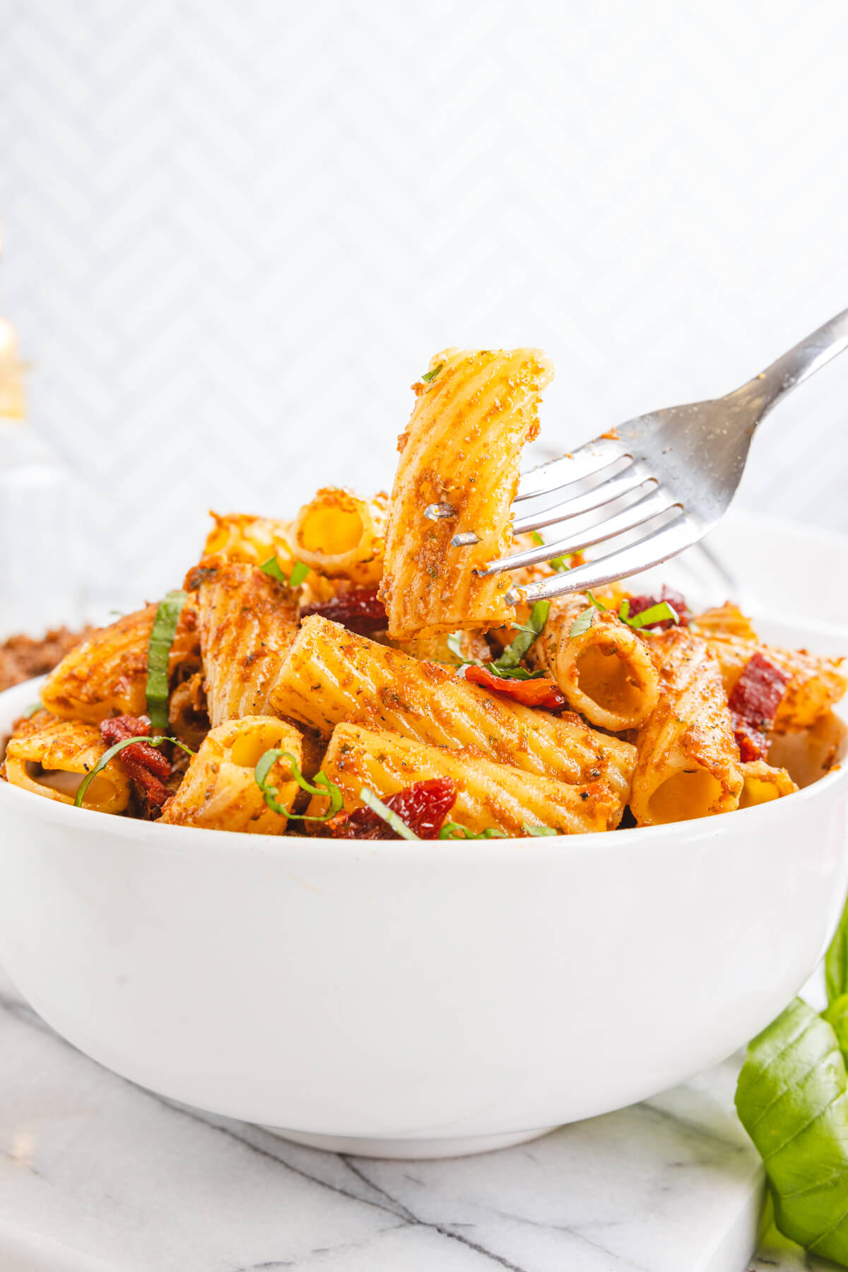 A fork grabbing a piece of rigatoni from a white bowl containing cooked rigatoni pasta coated in sundried tomato pesto garnished with fresh basil.