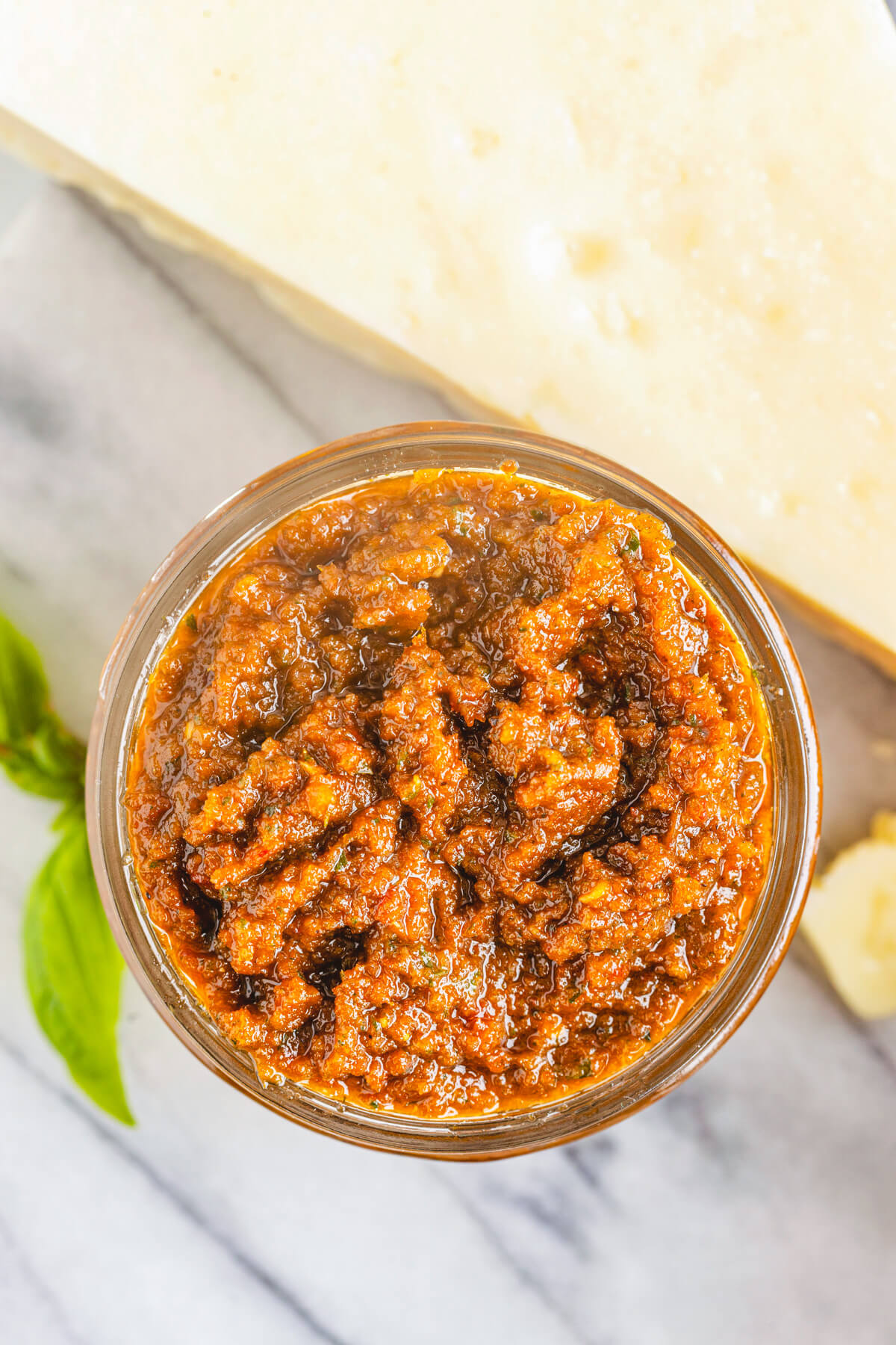 A jar of vibrant Sundried Tomato Pesto surrounded by fresh basil, Parmesan cheese, and garlic.