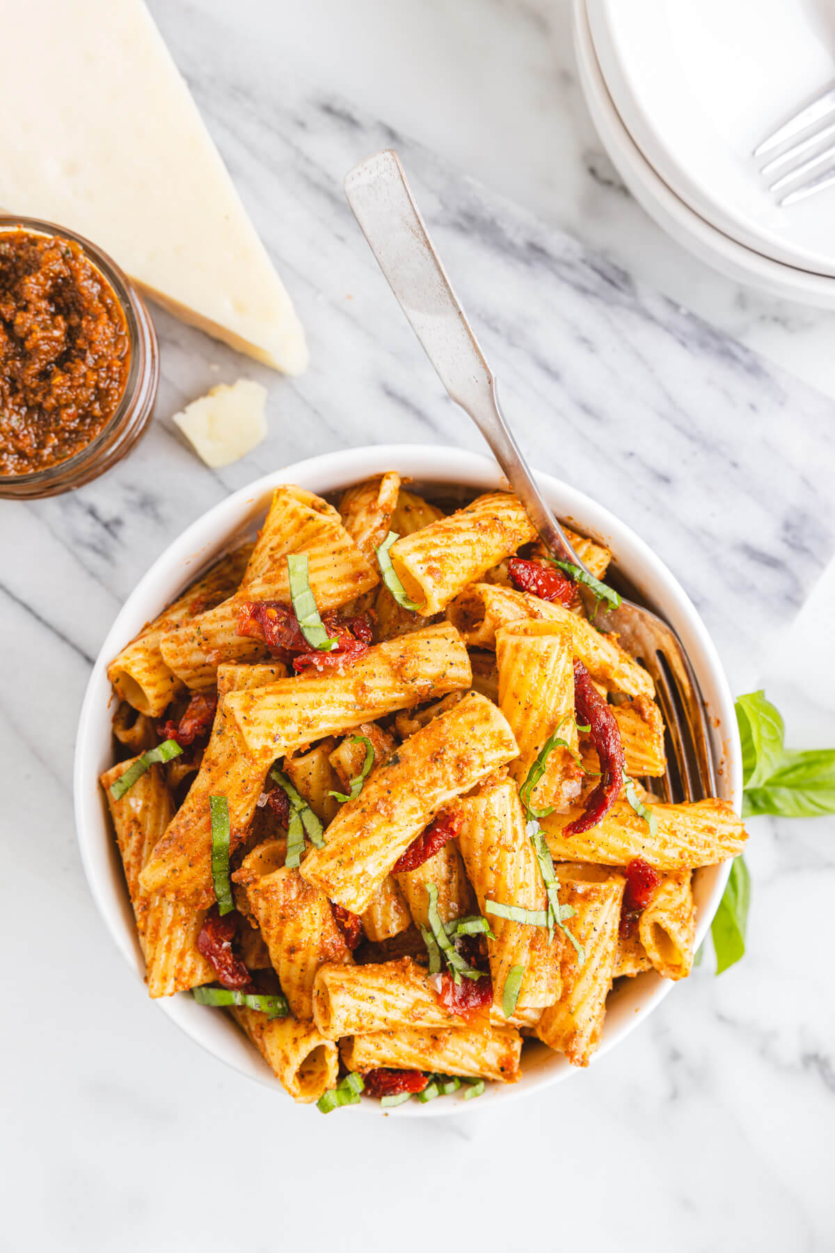 A white bowl containing cooked rigatoni pasta coated in sundried tomato pesto garnished with fresh basil.