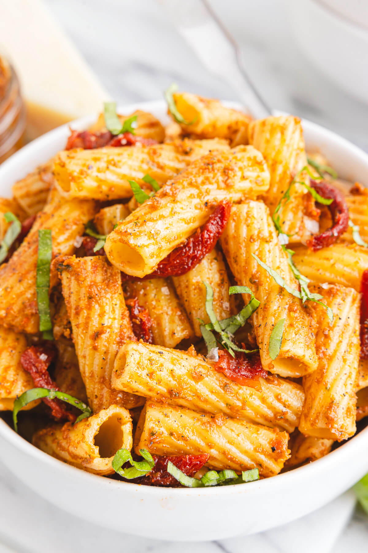 A white bowl containing cooked rigatoni pasta coated in sundried tomato pesto garnished with fresh basil.