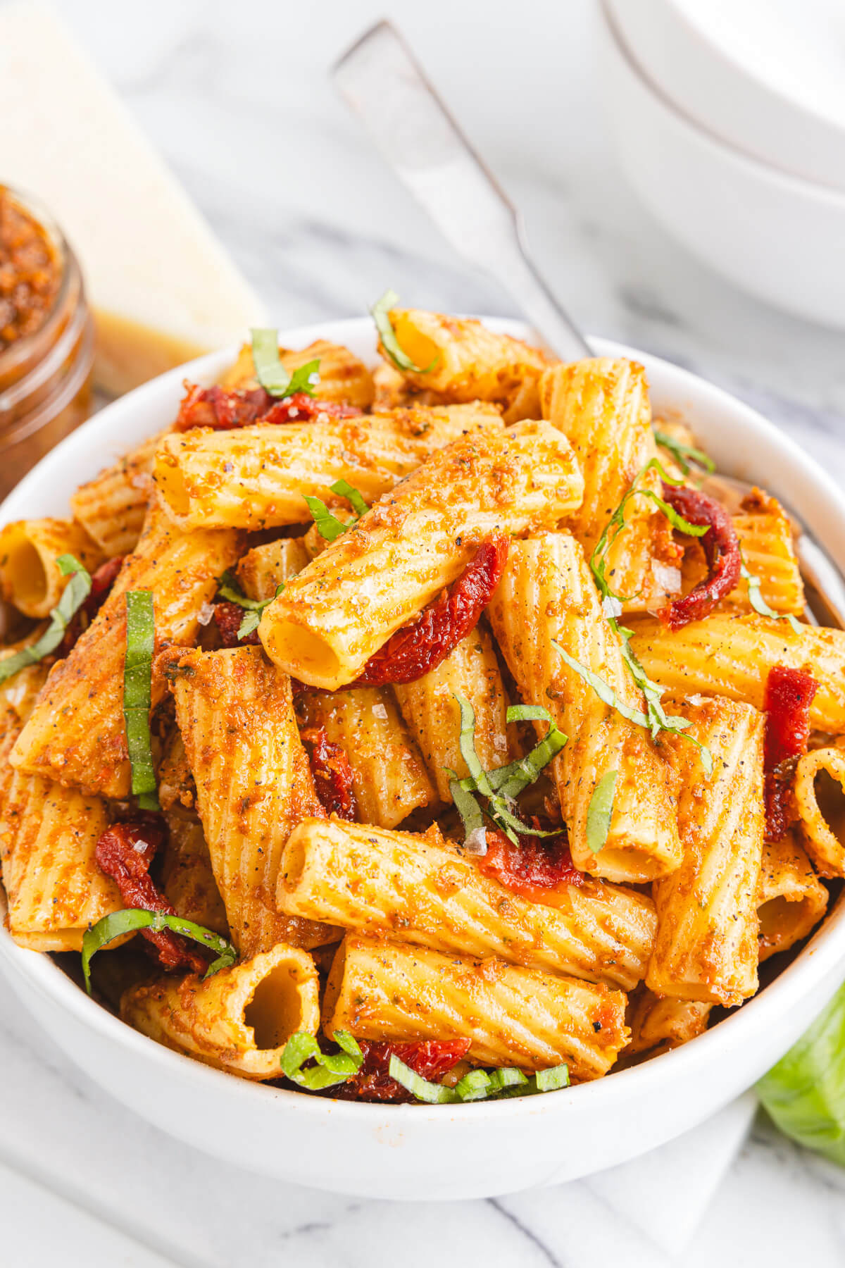 A white bowl containing cooked rigatoni pasta coated in sundried tomato pesto garnished with fresh basil.
