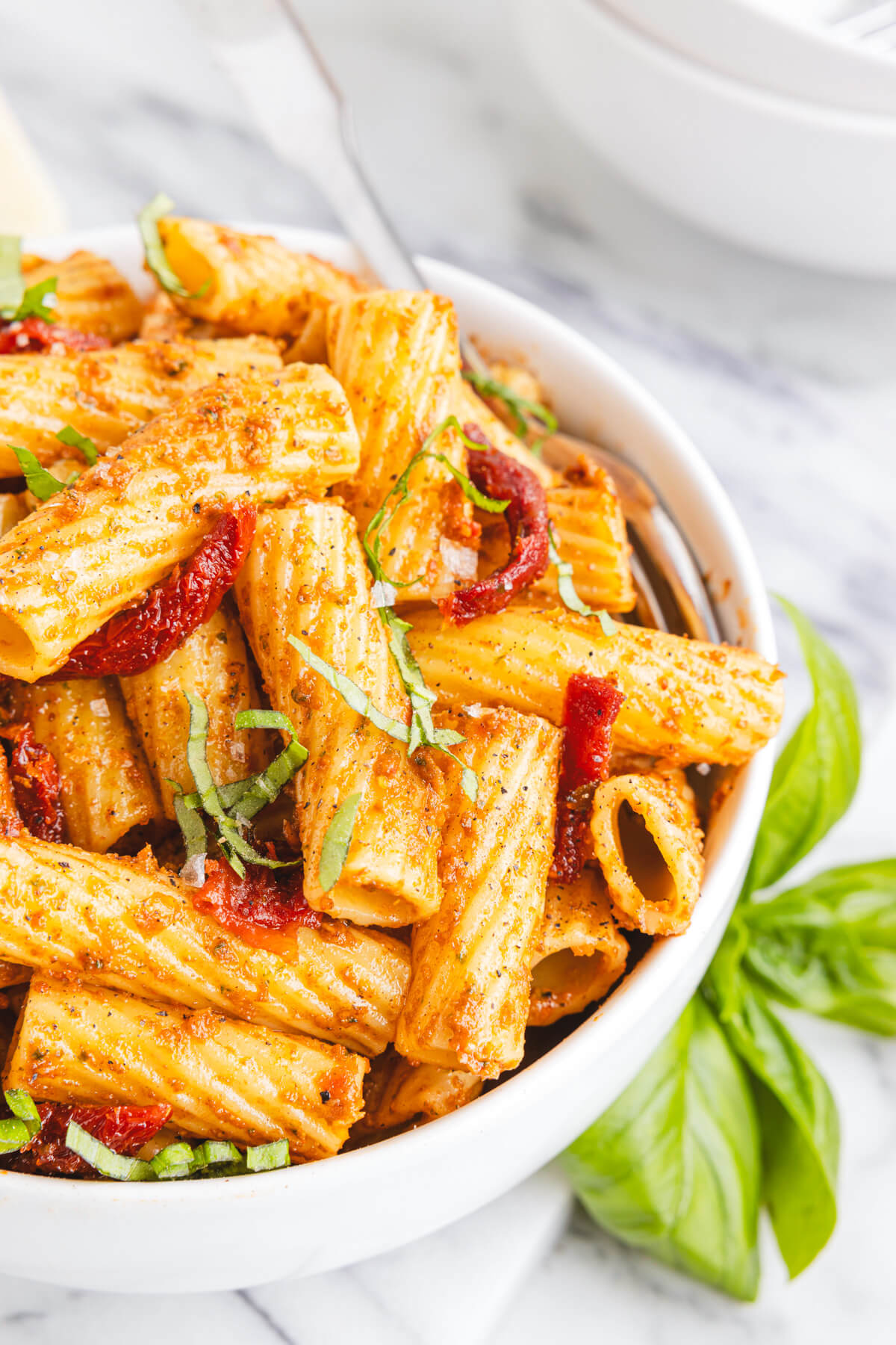 A white bowl containing cooked rigatoni pasta coated in sundried tomato pesto garnished with fresh basil.