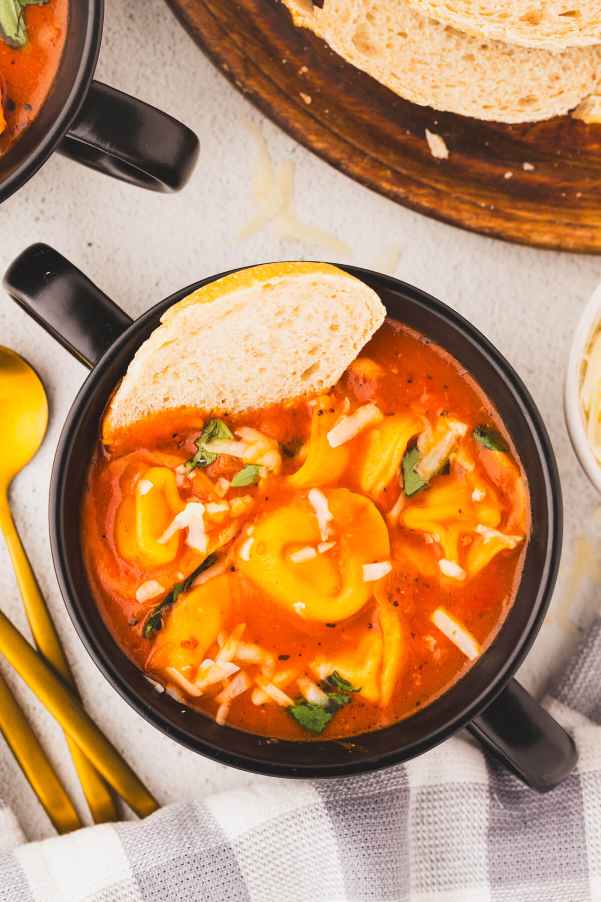 A black soup bowl with handles filled with creamy cheesy tomato tortellini soup and garnished with a slice of bread.