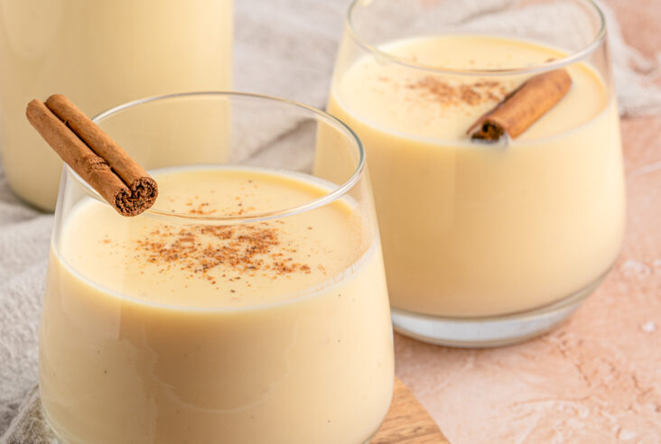 Two cocktail glasses of creamy Rompope, or Mexican Eggnog garnished with cinnamon sticks and ground cinnamon on a pink table.