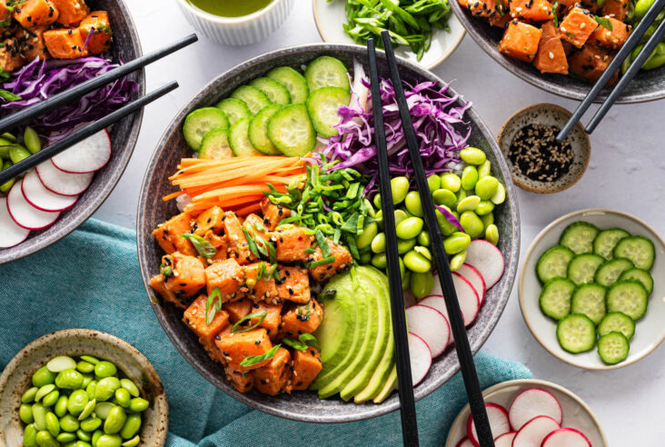 Black chopsticks sit on a vibrant Salmon Poke Bowl featuring green edamame, sliced avocado, julienned carrots, shredded purple cabbage, sliced radishes, and marinated diced salmon garnished with sesame seeds and sliced green onions.