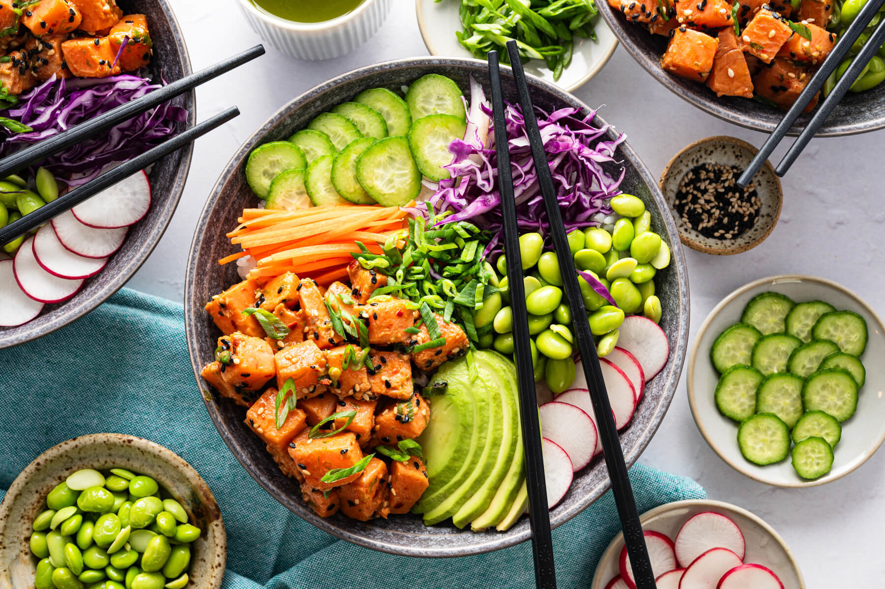 Black chopsticks sit on a vibrant Salmon Poke Bowl featuring green edamame, sliced avocado, julienned carrots, shredded purple cabbage, sliced radishes, and marinated diced salmon garnished with sesame seeds and sliced green onions.