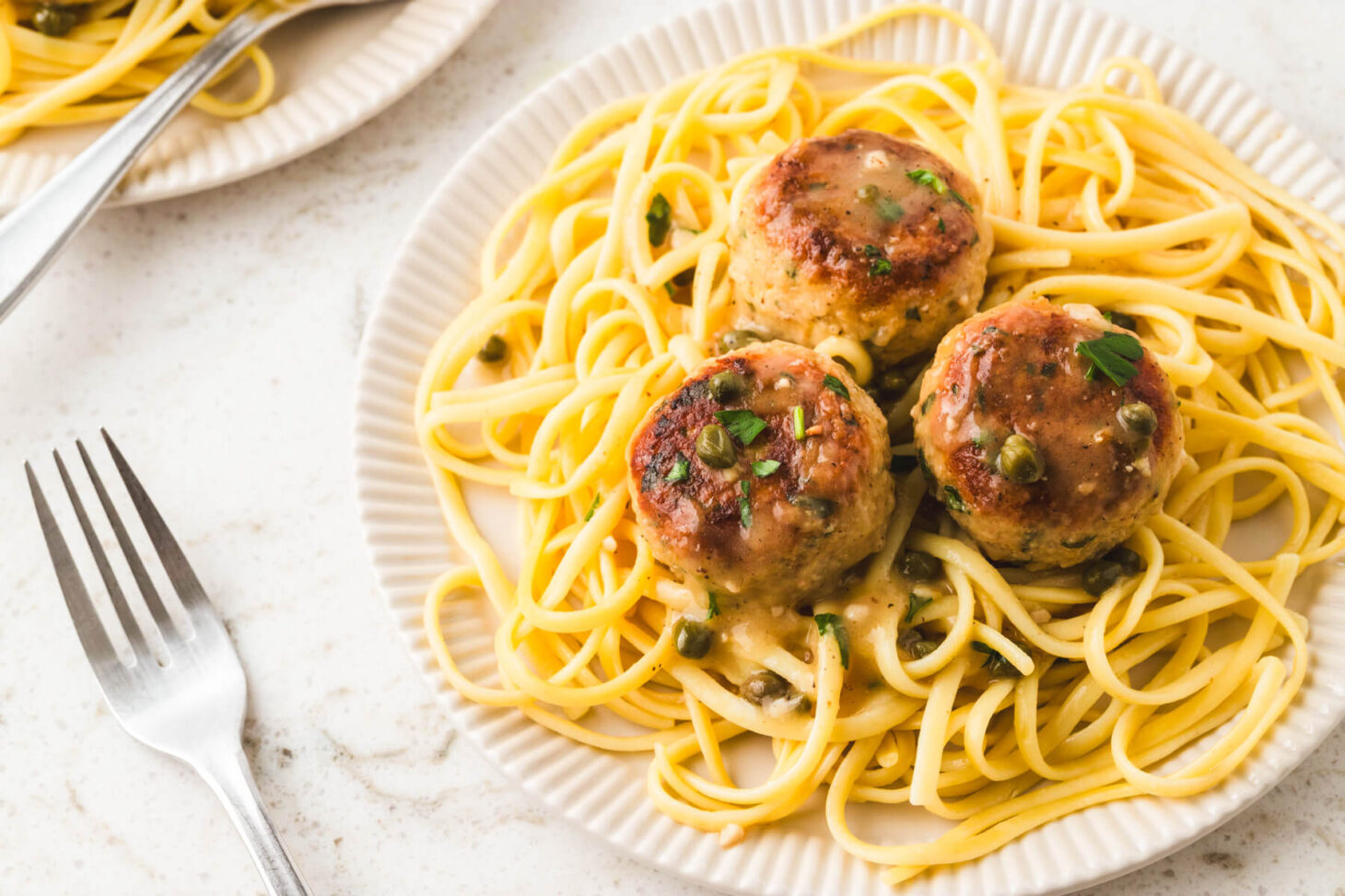 A white bowl containing chicken piccata meatballs on a bed of spaghetti. .