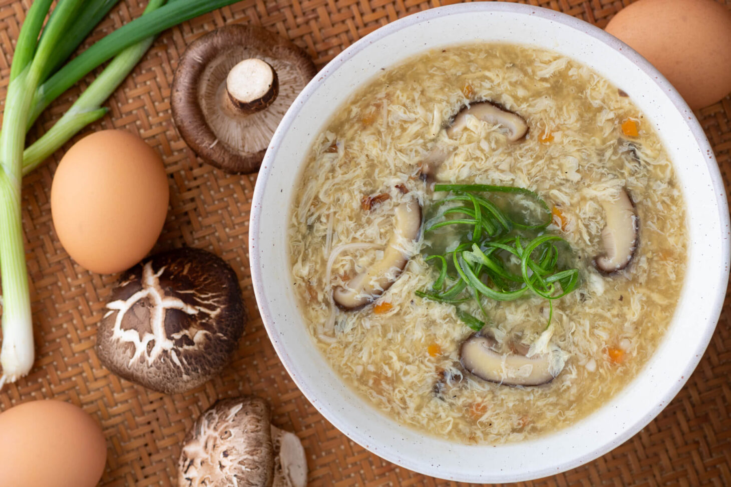 A white bowl of Egg Drop Soup containing shiitake mushrooms and finely diced carrots garnished with shredded green onions.