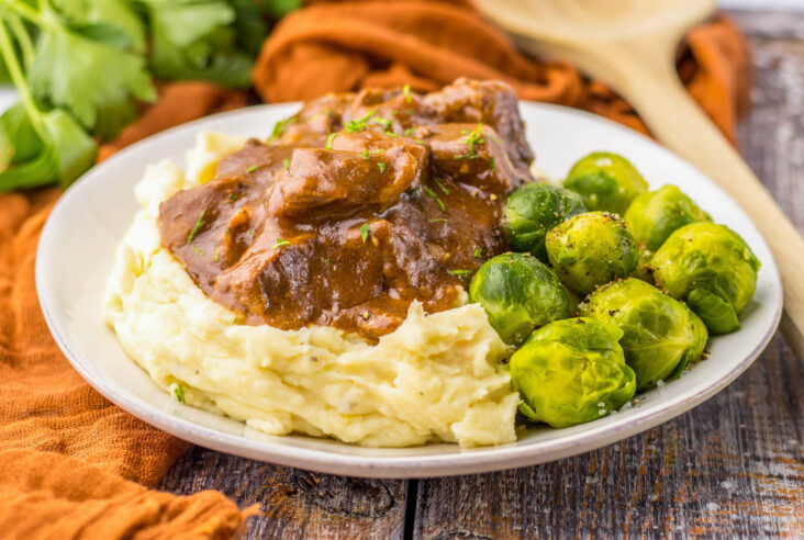 Tender slow cooked beef tips in a rich and flavourful gravy on a bed of mashed potatoes beside Brussels sprouts.