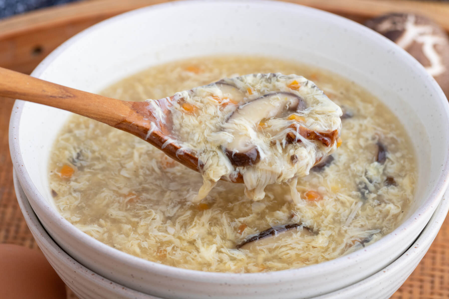 A wooden ladle filled with Egg Drop Soup hovers over a bowl of the same soup.