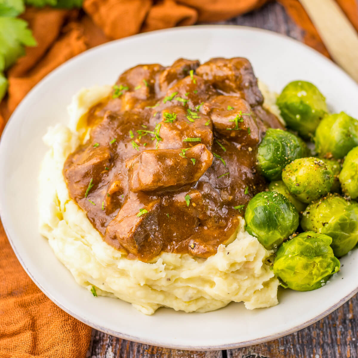 Tender slow cooked beef tips in a rich and flavourful gravy on a bed of mashed potatoes beside Brussels sprouts.