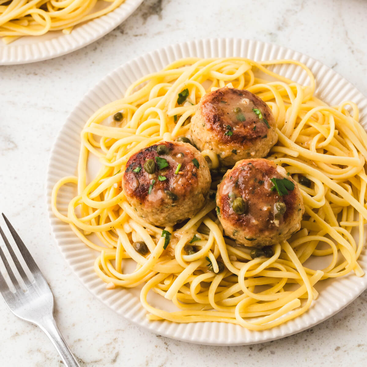 A white bowl containing chicken piccata meatballs on a bed of spaghetti. .