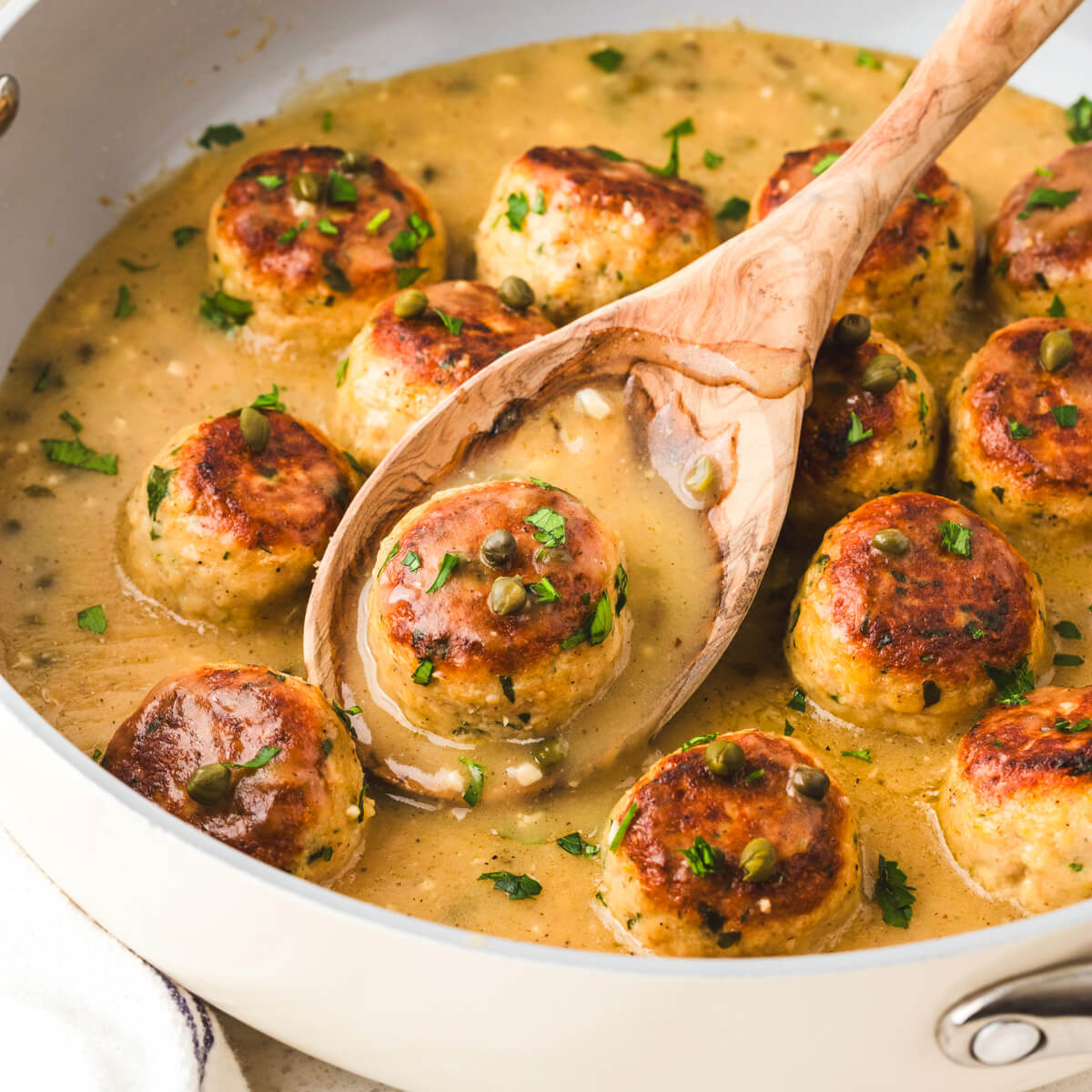 A pan full of cooked chicken meatballs in piccata sauce garnished with chopped Italian parsley.