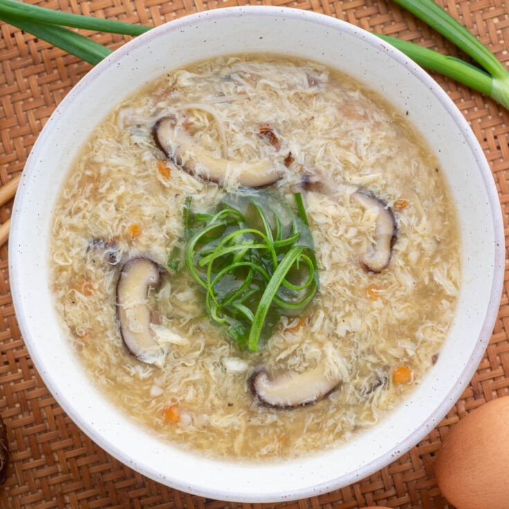 A white bowl of Egg Drop Soup containing shiitake mushrooms and finely diced carrots garnished with shredded green onions.