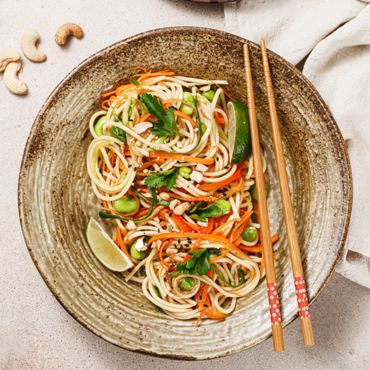 A bright and lively noodle salad featuring slivered carrots, red peppers, edamame, cucumbers, cashews, chopped cilantro, and lime wedges in a brown ceramic bowl with wooden chopsticks.