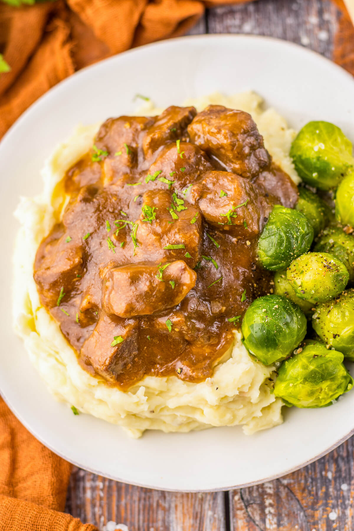 Tender slow cooked beef tips in a rich and flavourful gravy on a bed of mashed potatoes beside Brussels sprouts.