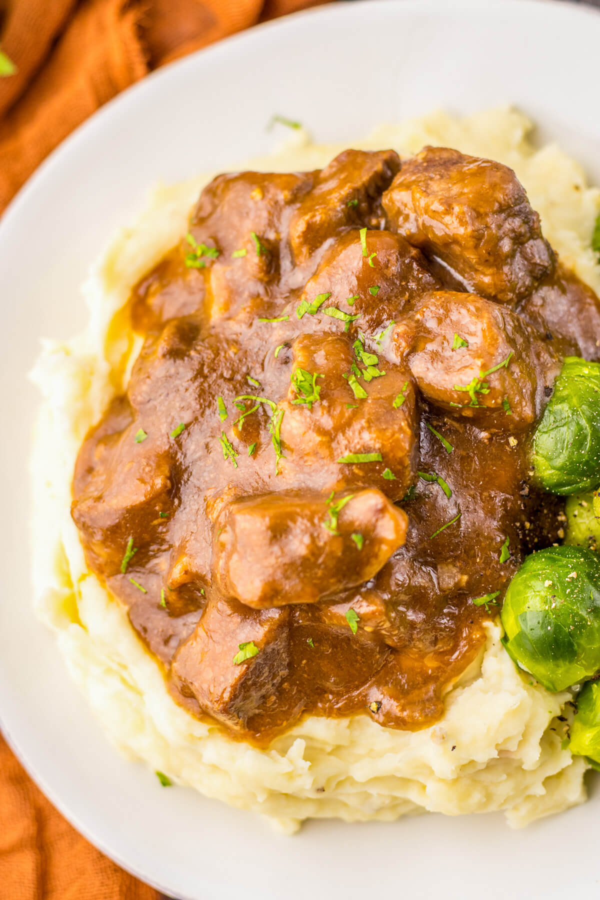Tender slow cooked beef tips in a rich and flavourful gravy on a bed of mashed potatoes beside Brussels sprouts.