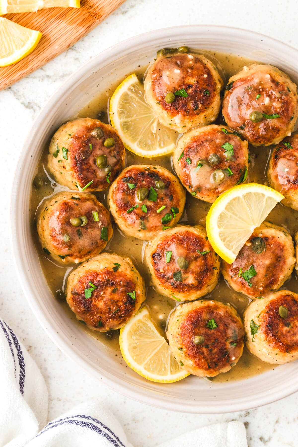 A white bowl containing chicken piccata meatballs garnished with wedges of lemon and chopped Italian parsley.