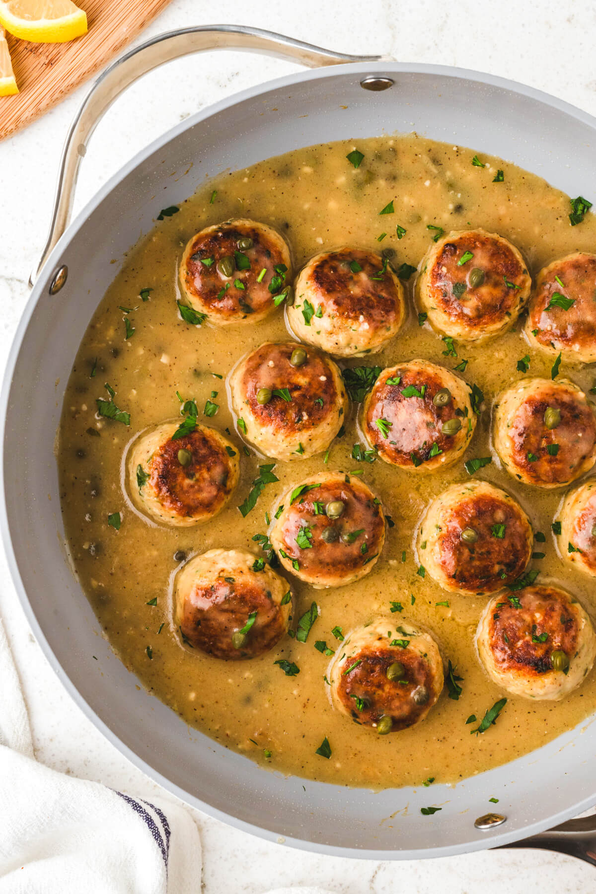 A pan full of cooked chicken meatballs in piccata sauce garnished with chopped Italian parsley.