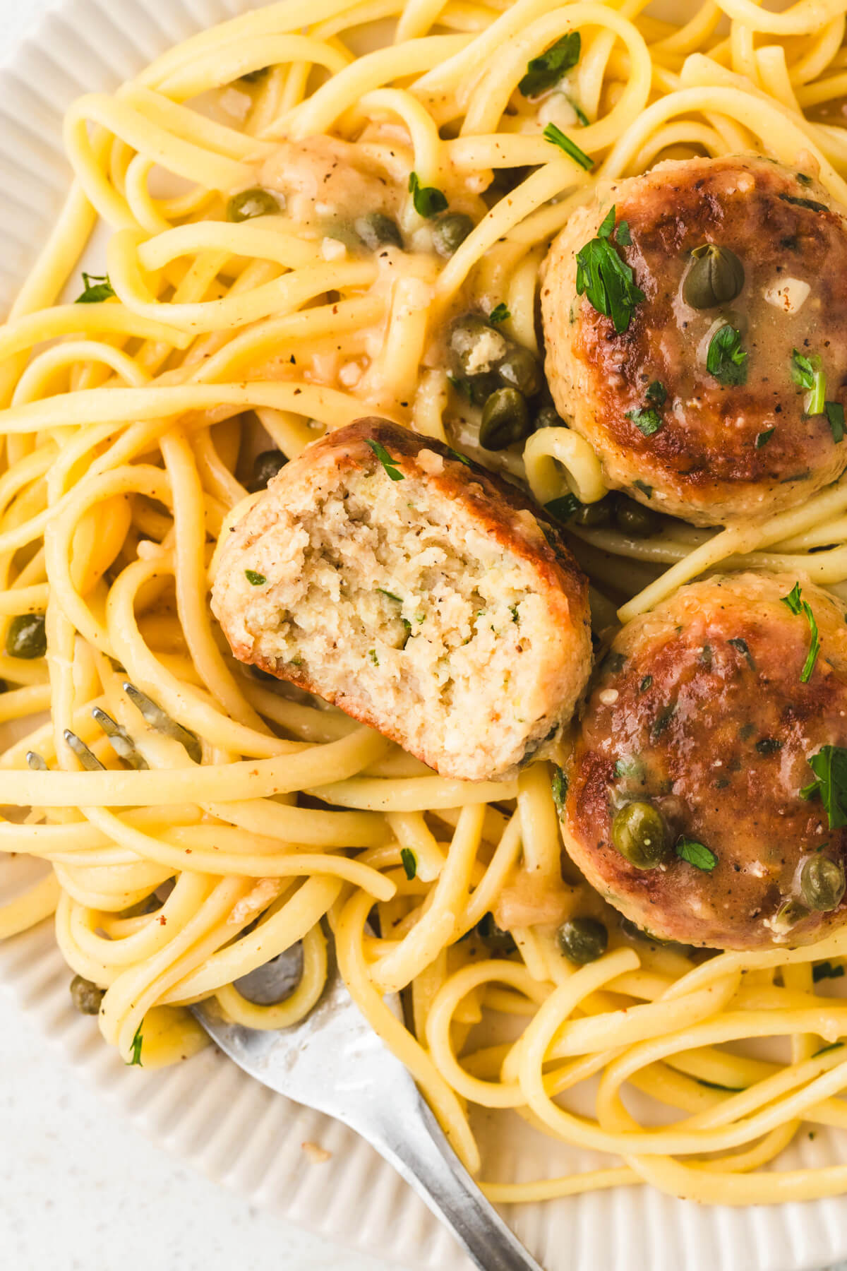 A white bowl containing three chicken piccata meatballs with one missing a bite on a bed of spaghetti. .