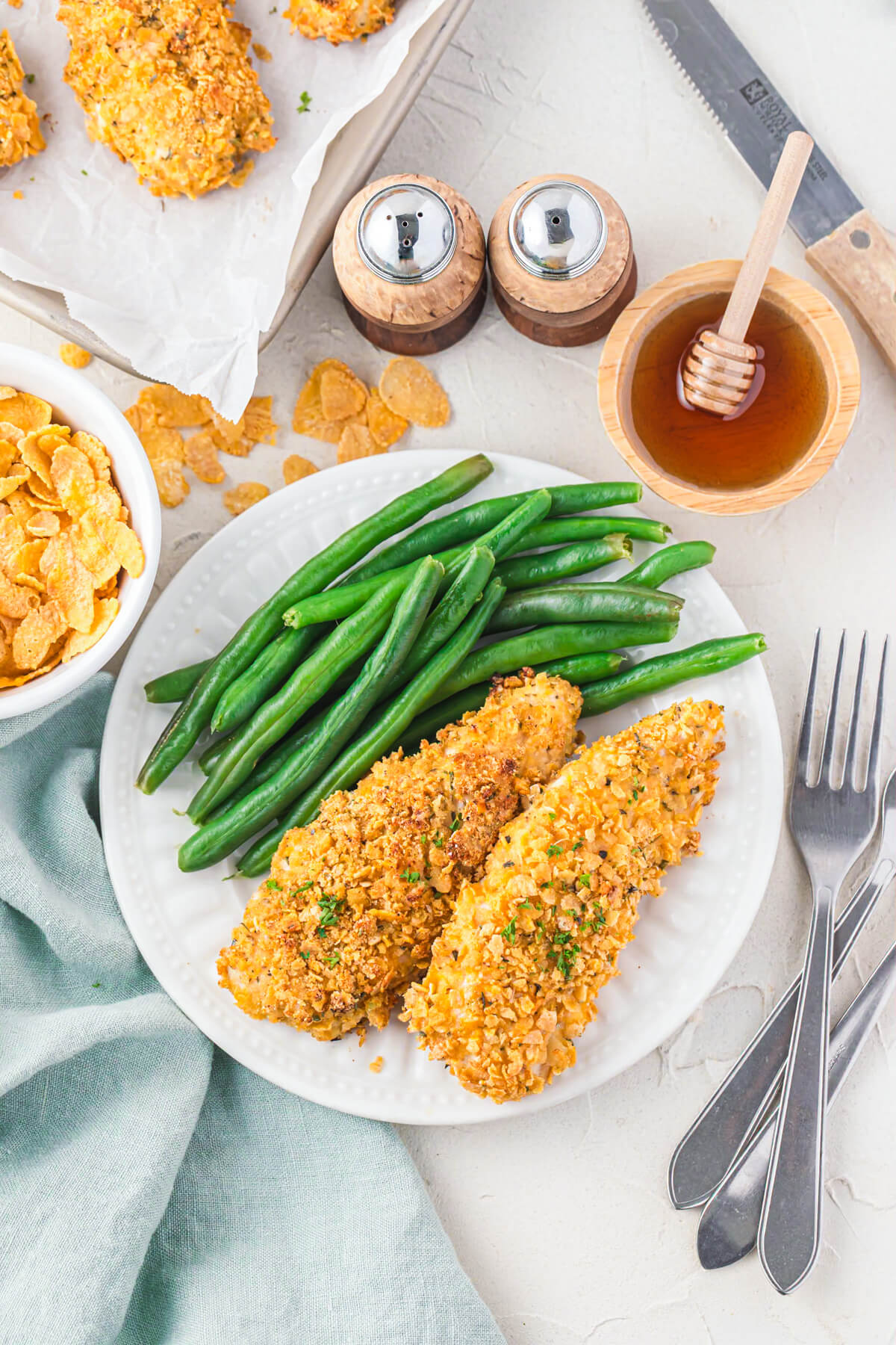 A white plate containing two golden cornflake chicken tenders and a side of steamed green beans on a table set for dinner.