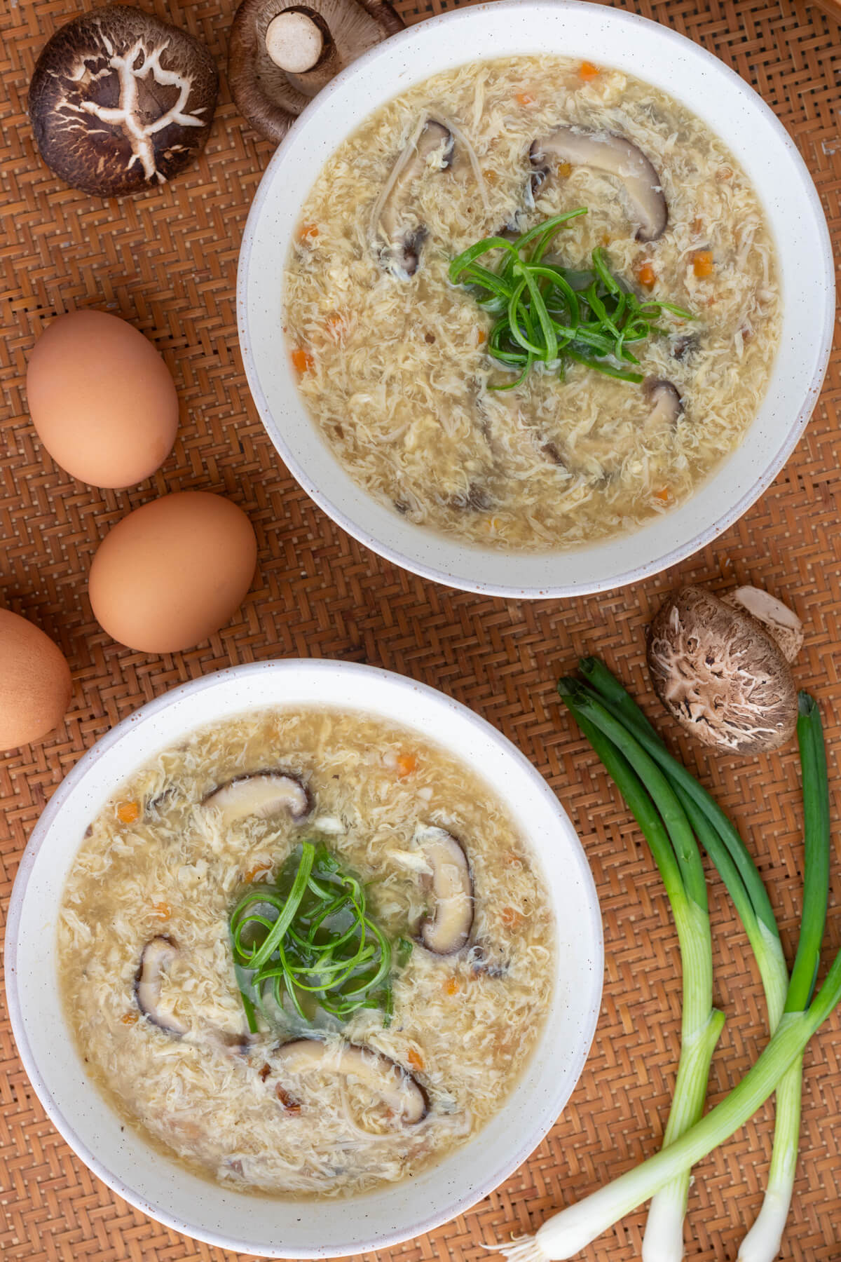 Two white bowls of Egg Drop Soup containing shiitake mushrooms and finely diced carrots garnished with shredded green onions.