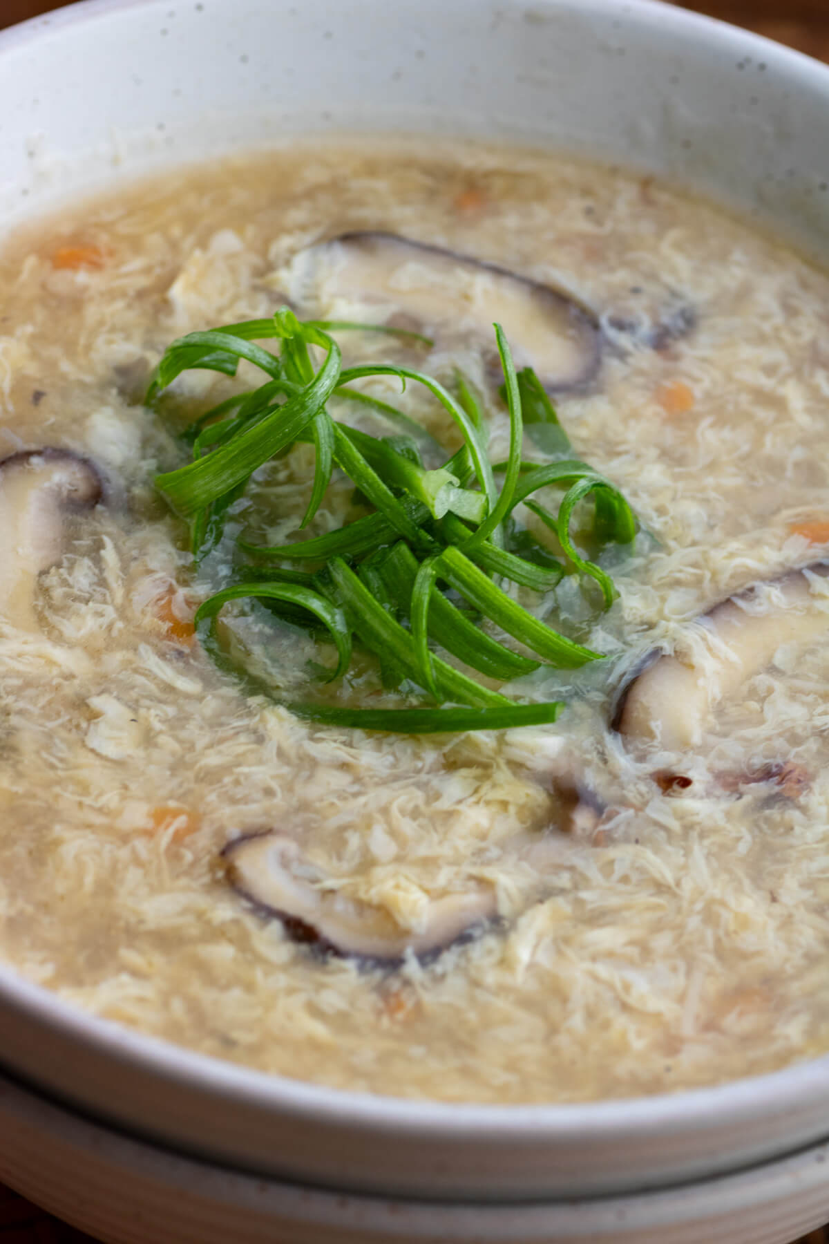 Close up view of a bowl of Egg Drop Soup containing shiitake mushrooms and finely diced carrots garnished with shredded green onions.