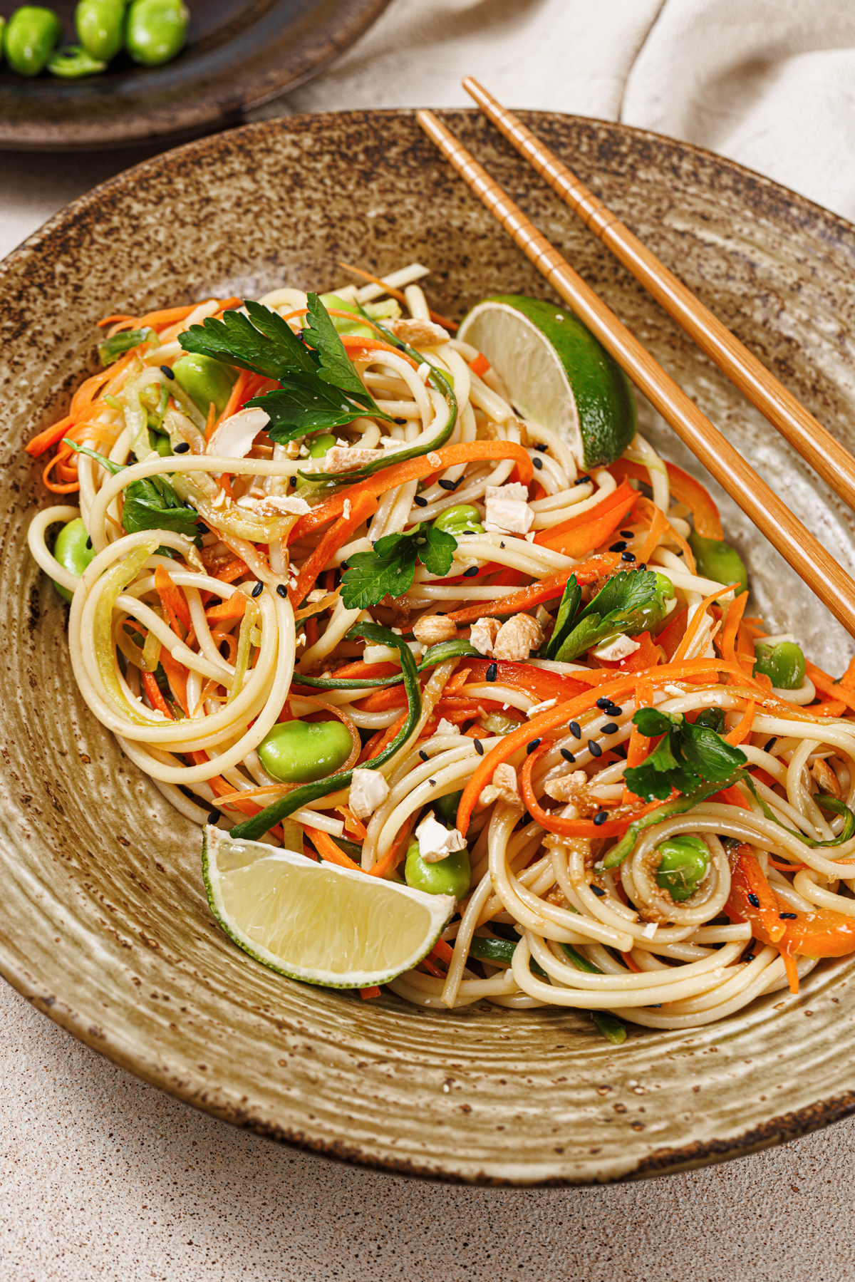 A bright and lively noodle salad featuring slivered carrots, red peppers, edamame, cucumbers, cashews, chopped cilantro, and lime wedges in a brown ceramic bowl with wooden chopsticks. 