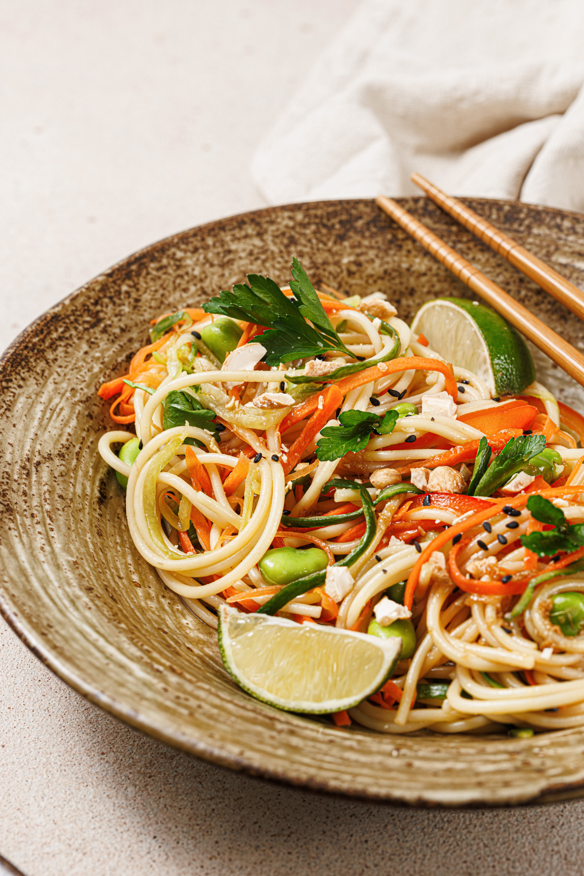 A bright and lively noodle salad featuring slivered carrots, red peppers, edamame, cucumbers, cashews, chopped cilantro, and lime wedges in a brown ceramic bowl with wooden chopsticks.