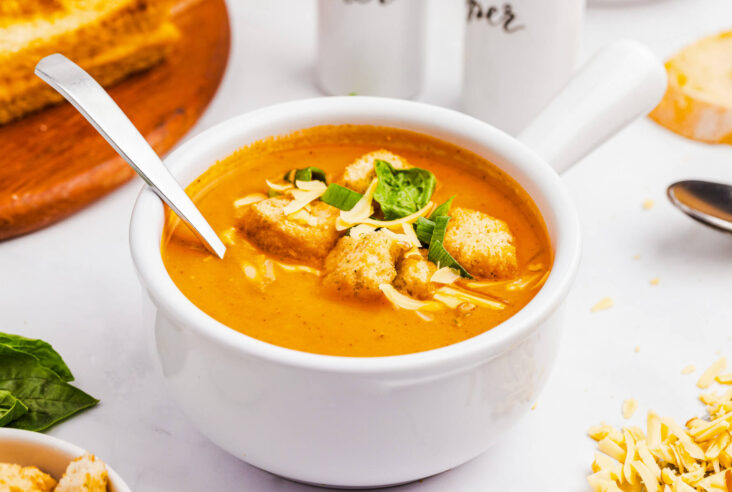 A bowl of bright orange, smooth and creamy Roasted Red Pepper Soup garnished with fresh basil, grated cheese and croutons.