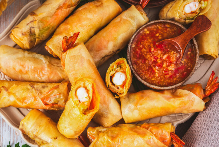 A party platter of golden fried shrimp spring rolls with tails sticking out the end of the wrappers next to a bowl of bright Thai chili sauce.