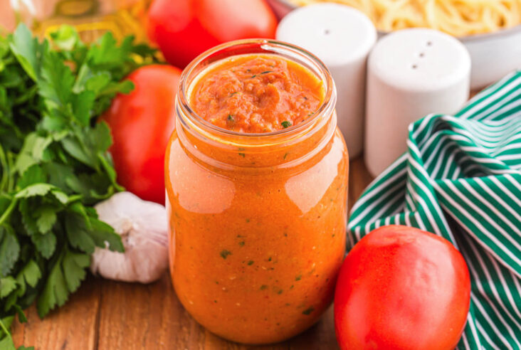 A jar of roasted tomato sauce nestled among sauce ingredients on a wooden table.