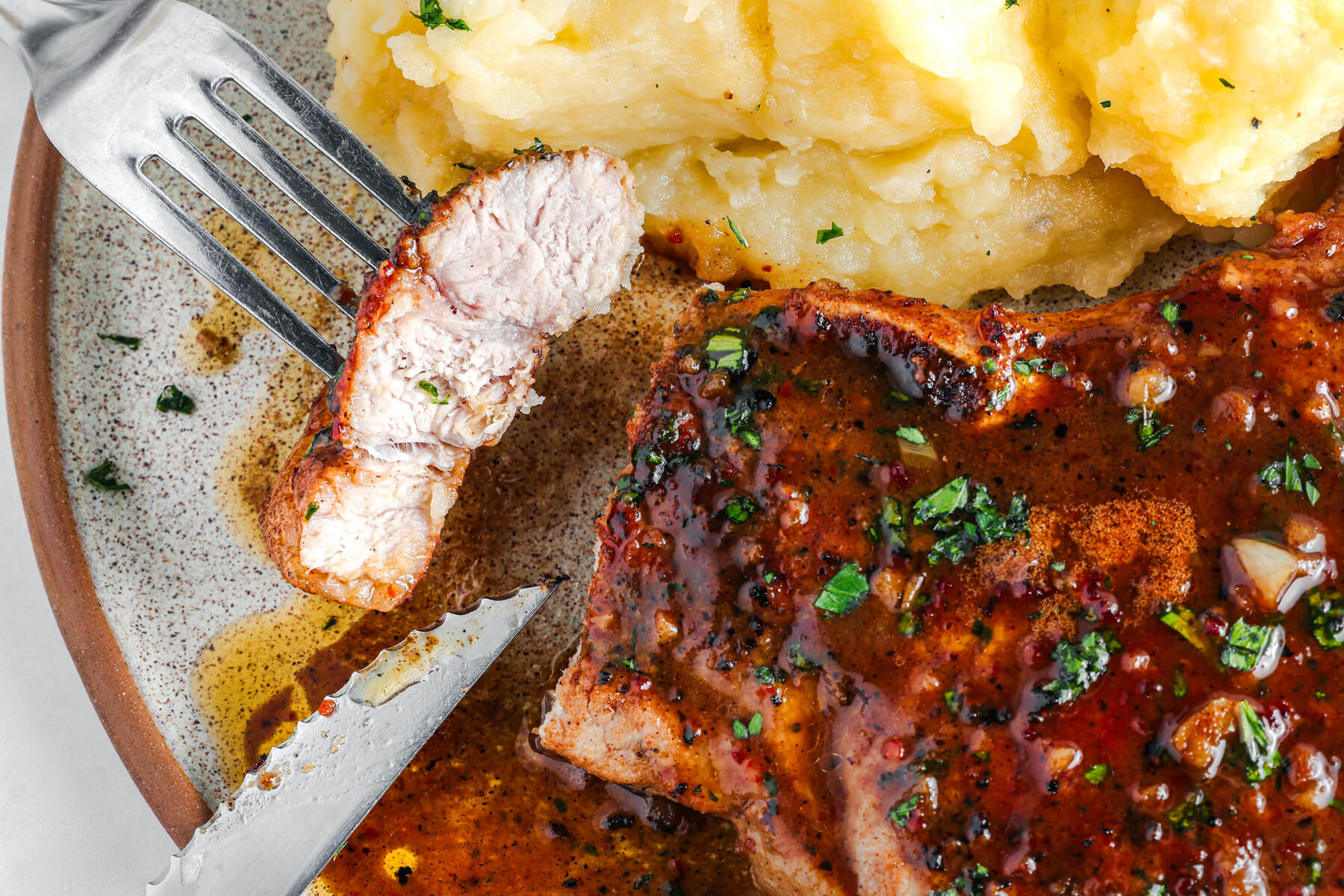 A fork holds a bite of pork chop while hovering over a dinner plate of mashed potatoes and Brown Sugar Pork Chops.