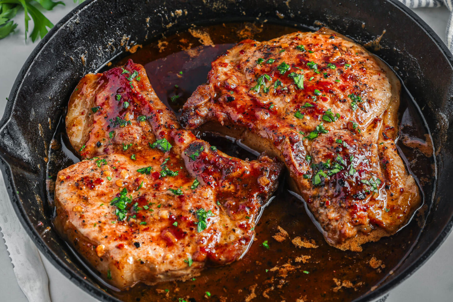 Two perfectly seared brown sugar pork chops covered in luxurious brown sauce, parsley, and garlic in a cast iron skillet.