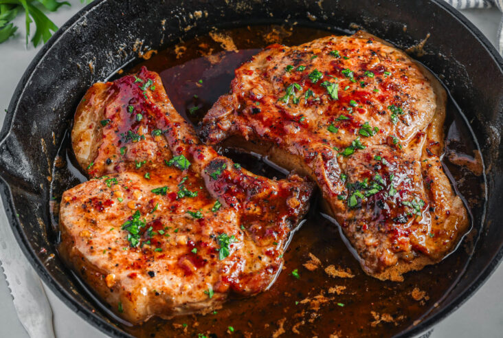 Two perfectly seared brown sugar pork chops covered in luxurious brown sauce, parsley, and garlic in a cast iron skillet.