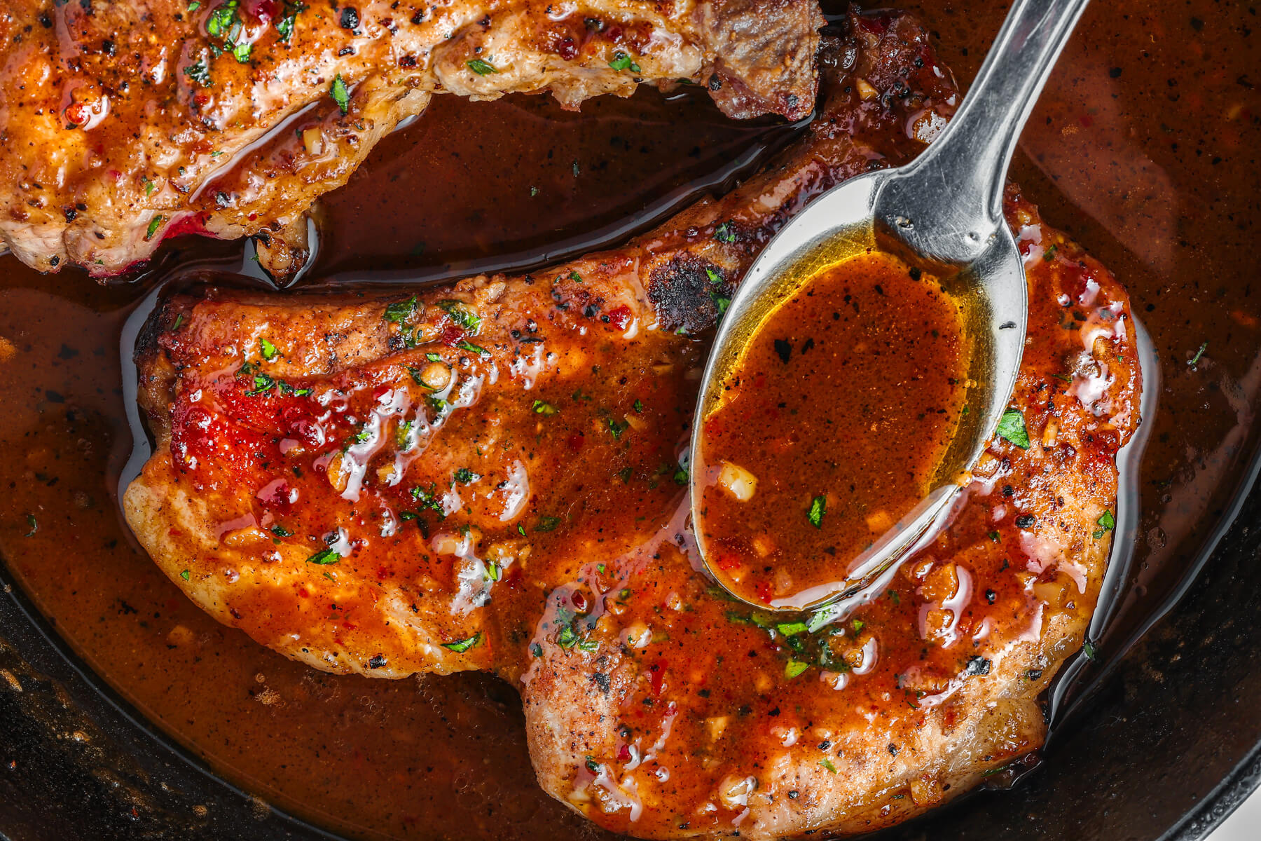 Two perfectly seared brown sugar pork chops being covered in luxurious brown sauce, parsley, and garlic in a cast iron skillet.