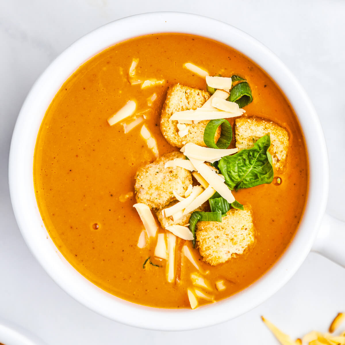 A bowl of bright orange, smooth and creamy Roasted Red Pepper Soup garnished with fresh basil, grated cheese and croutons.