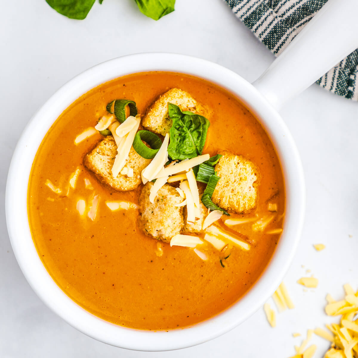 A bowl of bright orange, smooth and creamy Roasted Red Pepper Soup garnished with fresh basil, grated cheese and croutons.