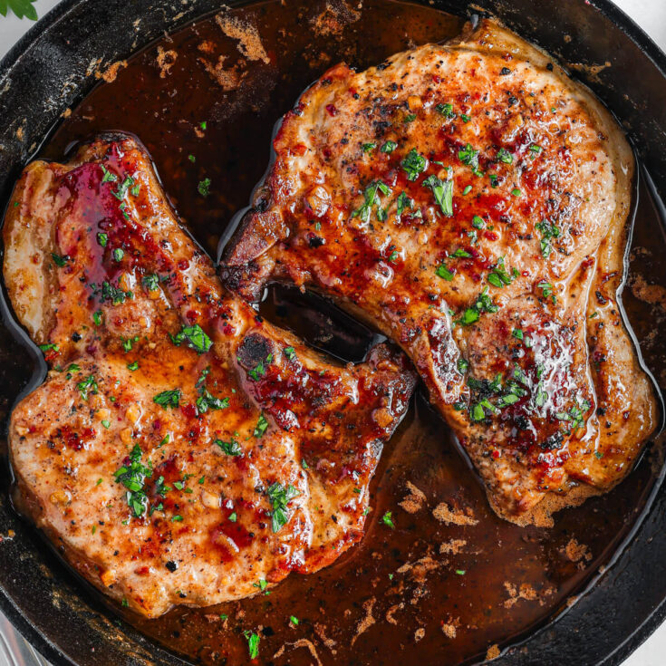 Two perfectly seared brown sugar pork chops covered in luxurious brown sauce, parsley, and garlic in a cast iron skillet.