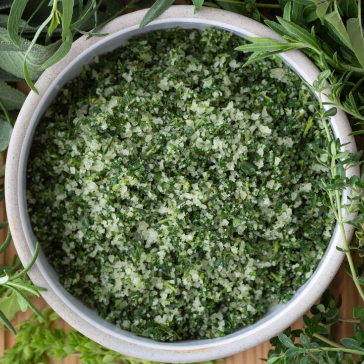 A round dish containing vibrant green herb salt surrounded by green herbs.