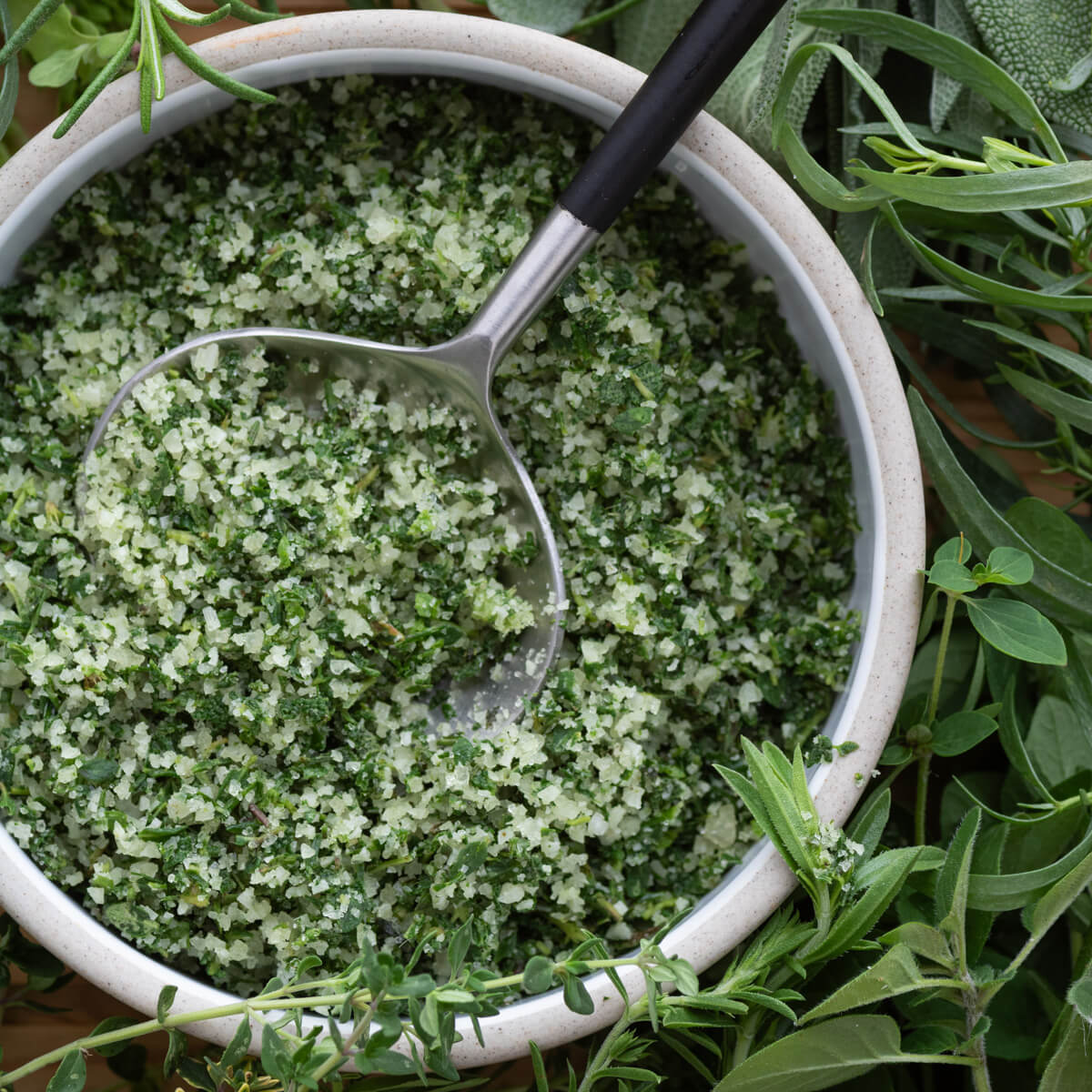A spoon resting in a round dish containing vibrant green herb salt surrounded by green herbs.