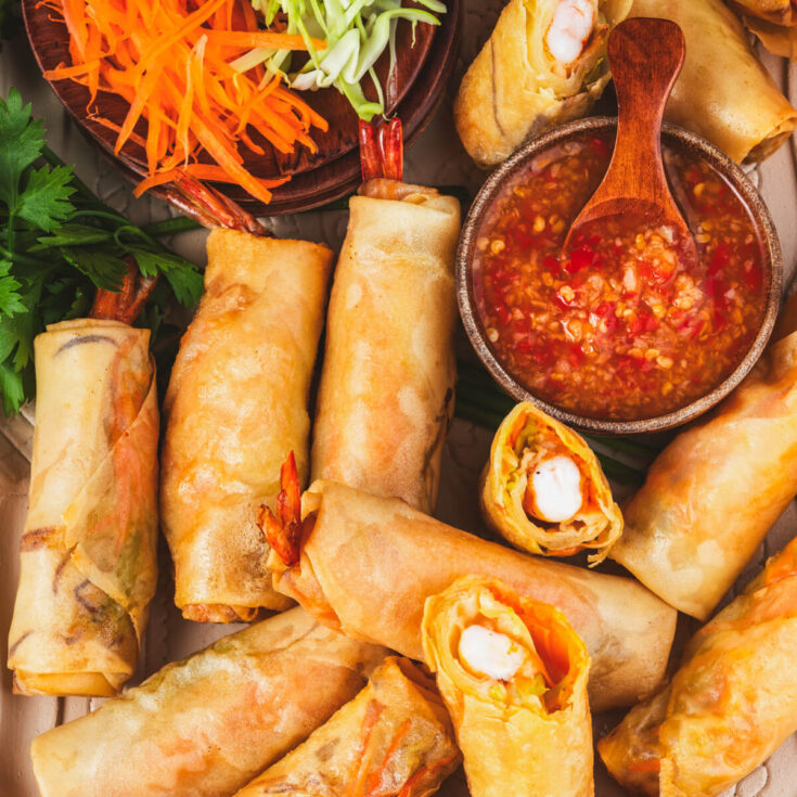 A party platter of golden fried shrimp spring rolls with tails sticking out the end of the wrappers next to a bowl of bright Thai chili sauce.
